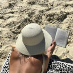 lady wearing a one piece bathing suit and a white straw hat laying on a towel on the sand, reading a book. Photo taken from behind.