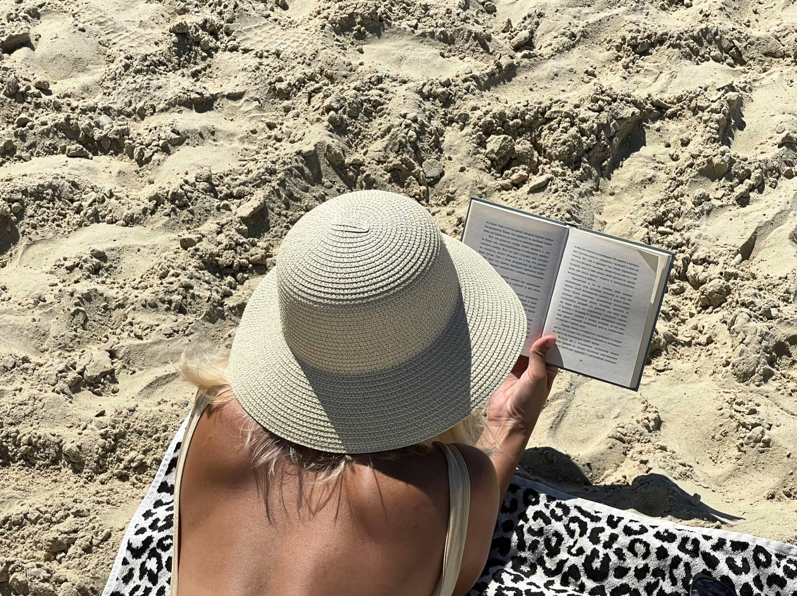 lady wearing a one piece bathing suit and a white straw hat laying on a towel on the sand, reading a book. Photo taken from behind.