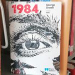 The cover of the Portuguese version of the book 1984 by George Orwell. The book is placed on a library shelf, facing the viewer, with a close up of a detailed realistic eye drawn in black ink, the title is bright red.